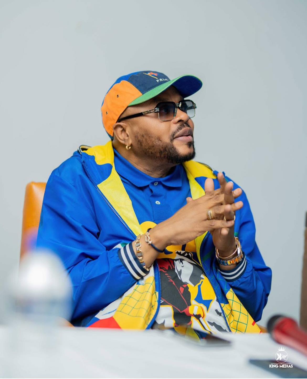 JB Mpiana, casquette bleue et orange, mains jointes, assis devant un micro